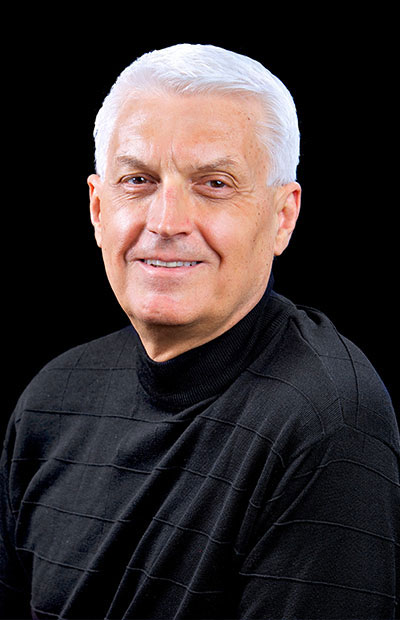 A man with gray hair and a beard, wearing a black shirt and smiling slightly, stands against a dark background.