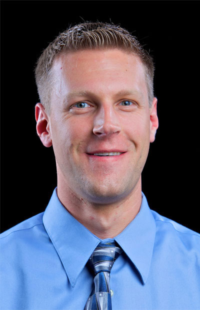 A man wearing a light blue shirt and tie, smiling directly at the camera.