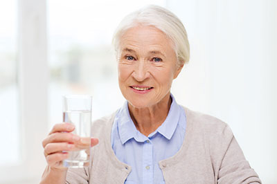 An elderly woman holding a glass of water, smiling at the camera.