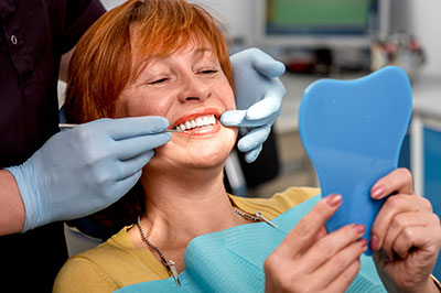 A woman sitting in a dental chair, smiling broadly, holding a blue toothbrush, with a dental hygienist assisting her.