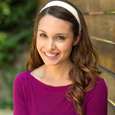 A young woman with long brown hair, wearing a purple top and a white headband, smiles against a wooden fence background.