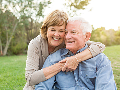 An elderly couple, a man and woman, embracing each other in an outdoor setting.