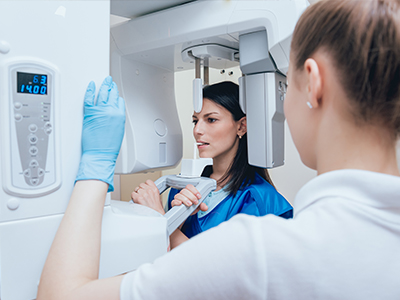 A woman in a blue coat stands before a large, modern CT scanner machine while a man in a white lab coat and gloves inspects the equipment.