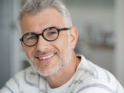 The image shows a man with glasses and a beard, smiling slightly at the camera. He has short hair and is wearing a white top with a patterned around the collar.