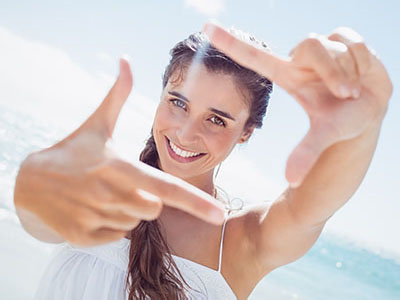 The image shows a smiling woman taking a selfie with her hand, set against a sunny beach backdrop.