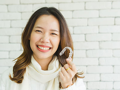 A smiling woman holding a clear aligner tray with her mouth, set against a brick wall background.